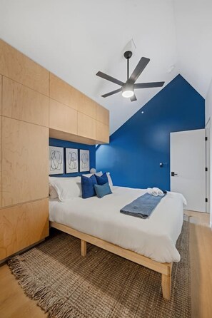 A modern bedroom with a sloped ceiling, a bed with white bedding, blue pillows, and a ceiling fan. - 3-Story Designer Townhome by City Park (Manhattan)