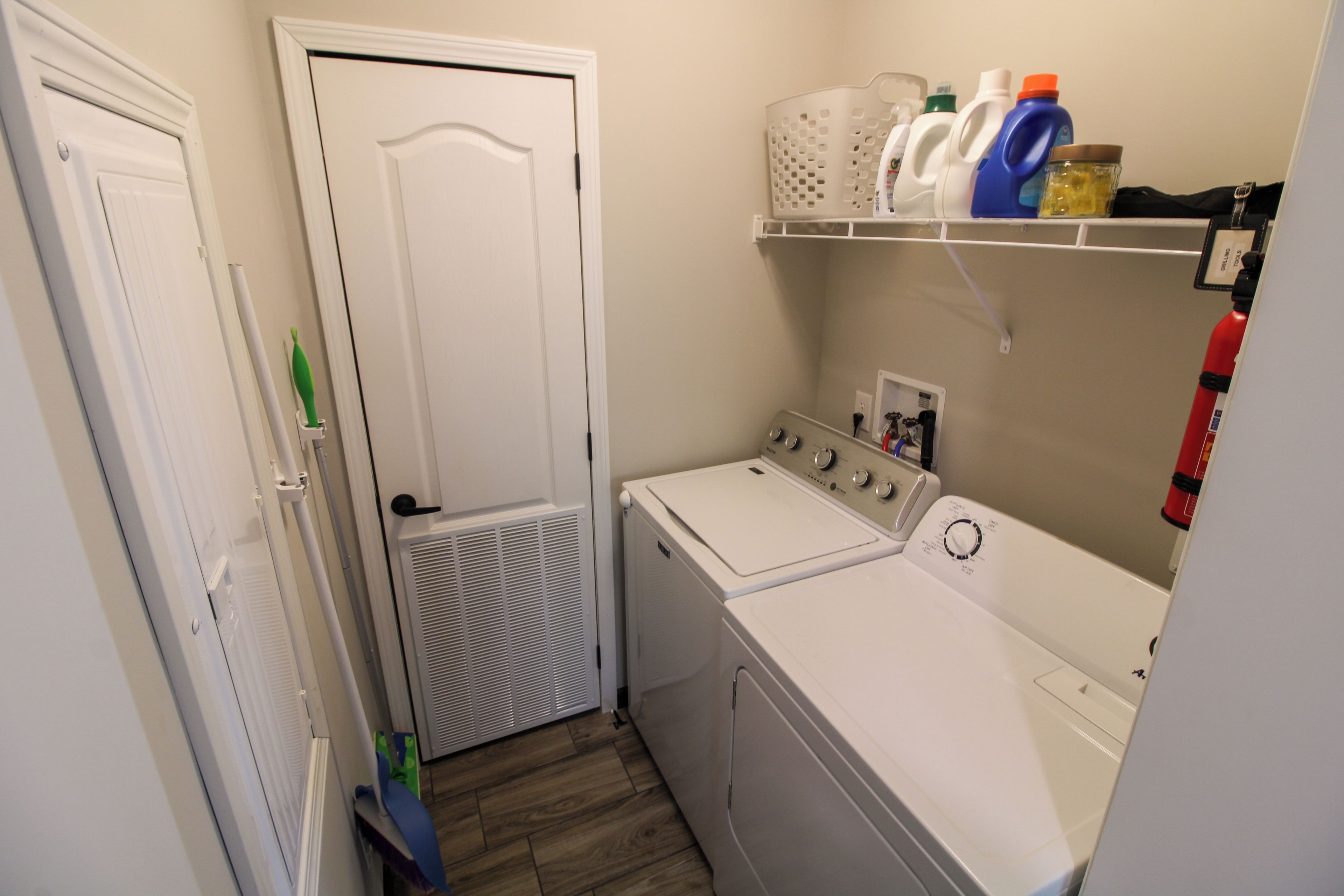 A laundry room with a washer and dryer, a shelf with cleaning supplies, and a door leading to another room.