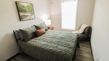 Quarto com cama, mesa de cabeceira, abajur, cadeira e janela.