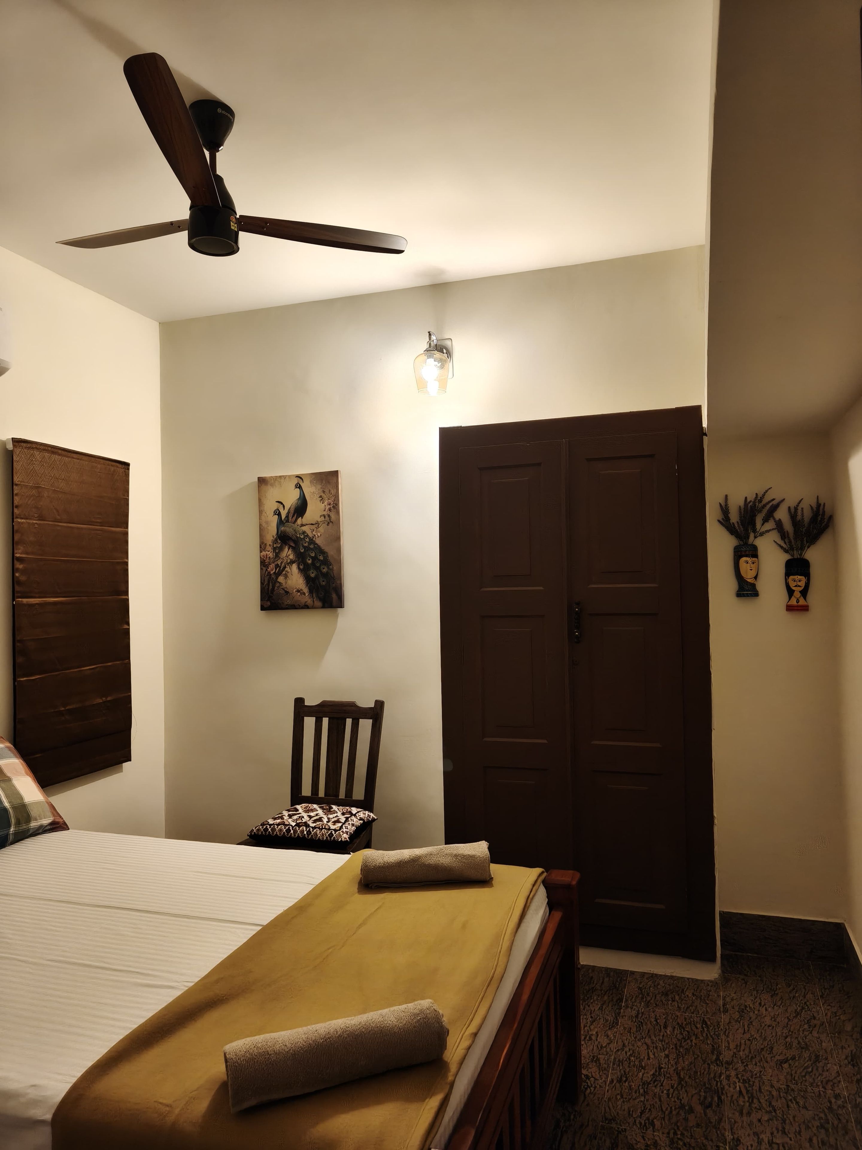 A hotel room with a bed, a chair, a ceiling fan, a door, and a wall-mounted light fixture.