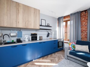 A modern kitchen with blue cabinets, a microwave, a kettle, and a coffee machine. There is a brick wall and a window with curtains. - Wółczańska Central Studio (Łódź)