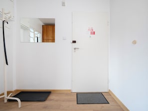 A room with a white door, a black mat at the entrance, a wooden floor, a white wall, and a mirror reflecting a wooden cabinet. - Maratońska Standard Apartment (Łódź)