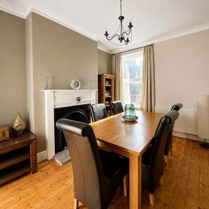 A dining room with a wooden table and black chairs, a fireplace, a bookshelf, and a window with curtains. - Cosy Victorian Home in Levenshulme's Trendy Suburb (Manchester)