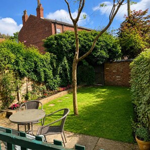 A backyard with a brick house, a grassy lawn, a tree, and a patio with a table and chairs. - Cosy Victorian Home in Levenshulme's Trendy Suburb (Manchester)