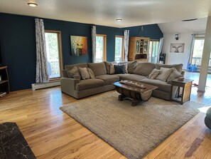 A living room with a sectional sofa, a coffee table, and a rug. There are windows with curtains, a painting on the wall, and a wooden cabinet. - 4 Bedroom Private Mountain Home Near Golden, Boulder, Eldora Ski Resort (Golden)