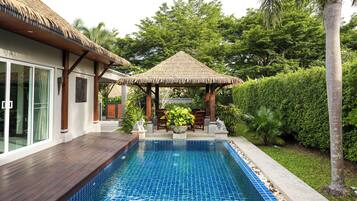 Une piscine agrémentée d’un gazebo à toit de chaume, entourée d’une végétation luxuriante et d’une maison moderne dotée de grandes fenêtres.