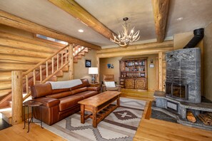 A log cabin interior with a wooden staircase, leather sofa, fireplace, and a bookcase. - NEW | Private Hot Tub | Hike | Gallatin Canyon, Fire Pit, Sleeps 12 (Gallatin Gateway)