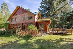 A log cabin with a green roof, surrounded by dense forest. - NEW | Private Hot Tub | Hike | Gallatin Canyon, Fire Pit, Sleeps 12 (Gallatin Gateway)