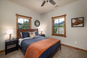 A bedroom with a bed, two nightstands, a ceiling fan, and two windows with views of mountains. - New! Private Hot Tub | Town Center | Sleeps 9 (Big Sky)