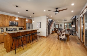 A modern kitchen with wooden cabinets, a central island with bar stools, and a dining area with a table and chairs. - New! Private Hot Tub | Town Center | Sleeps 9 (Big Sky)