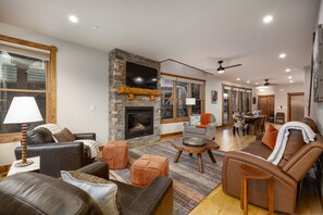 A spacious living room with a stone fireplace, leather sofas, a dining area, and a large window. - New! Private Hot Tub | Town Center | Sleeps 9 (Big Sky)