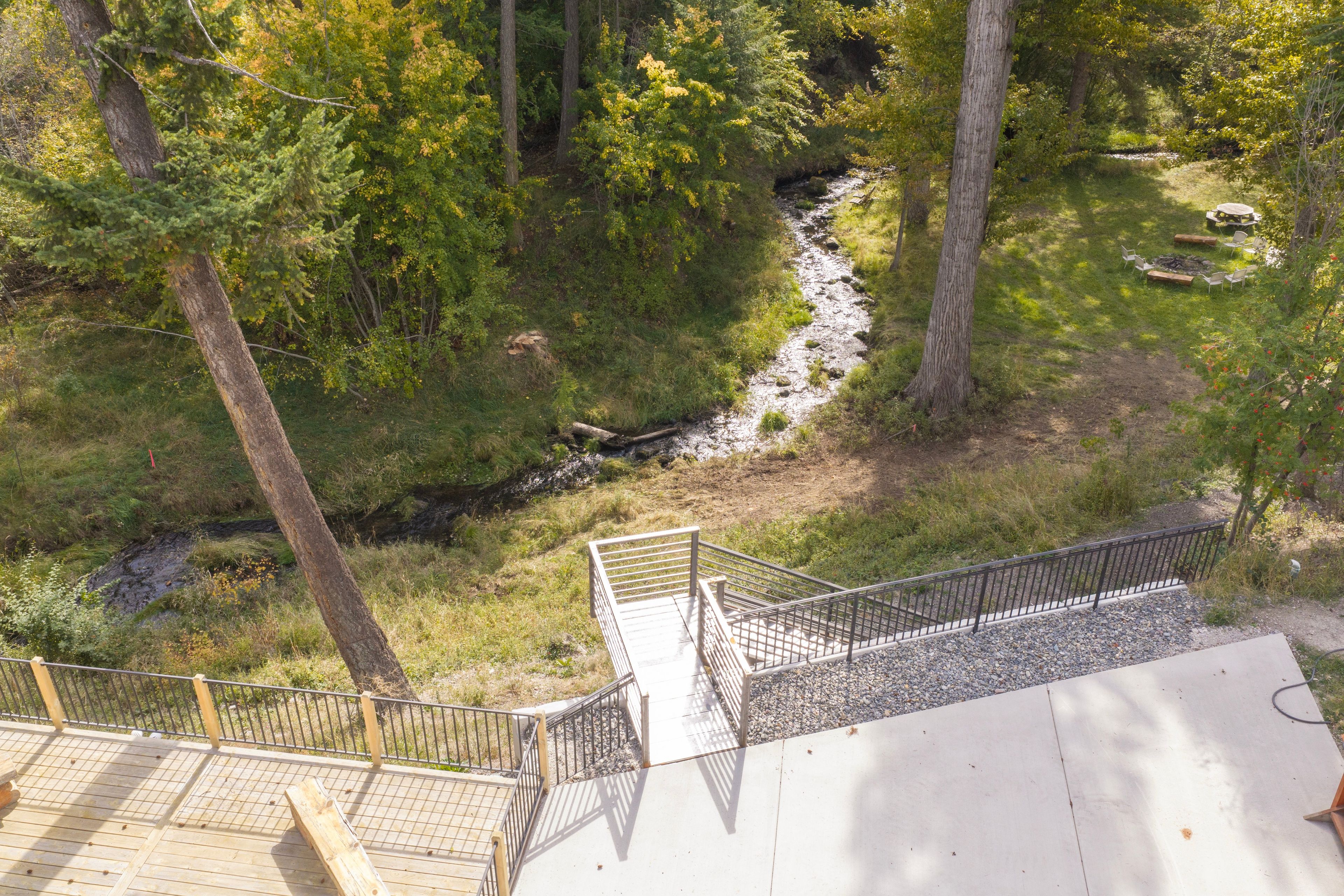 Une terrasse en bois avec une rambarde en métal, donnant sur une zone boisée traversée par un ruisseau.