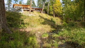 Une cabane dotée d’une terrasse, entourée de forêt et bordée par un ruisseau.