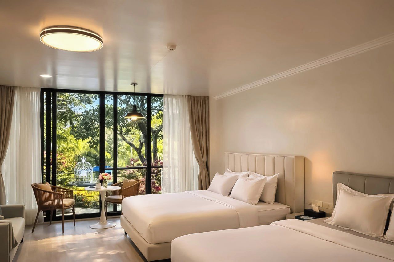 A hotel room with two beds, a dining area with a table and chairs, and large windows with a view of greenery.