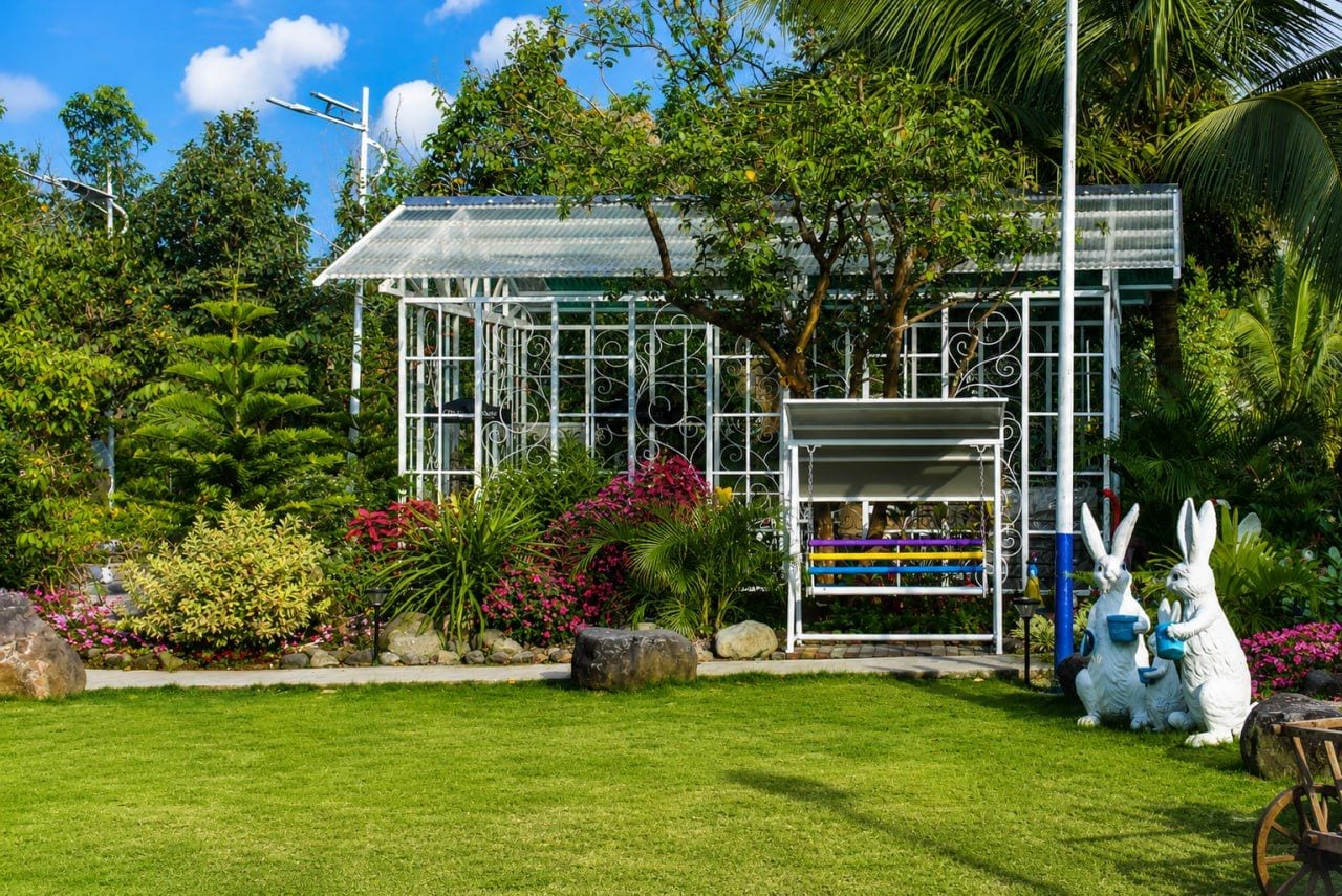 A greenhouse with a metal frame, surrounded by lush greenery and flowering plants.