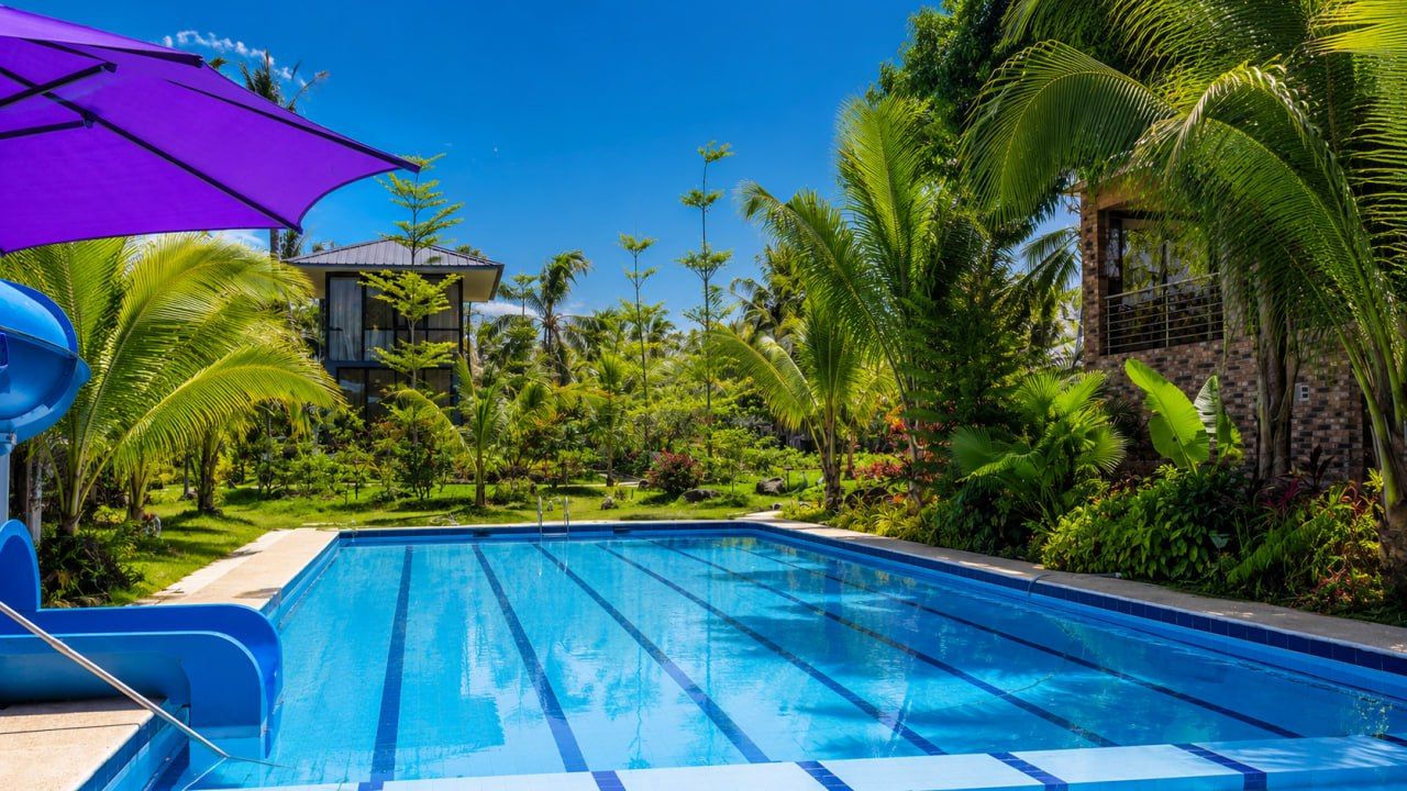 A swimming pool with clear blue water, surrounded by lush greenery and a purple umbrella.