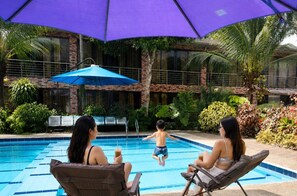 Una piscina exterior con una sombrilla azul, rodeada de exuberante vegetación y un edificio de ladrillo.
