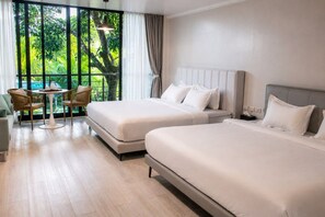 Habitación de hotel con dos camas, una pequeña mesa con sillas y vista a zonas verdes a través de amplios ventanales.