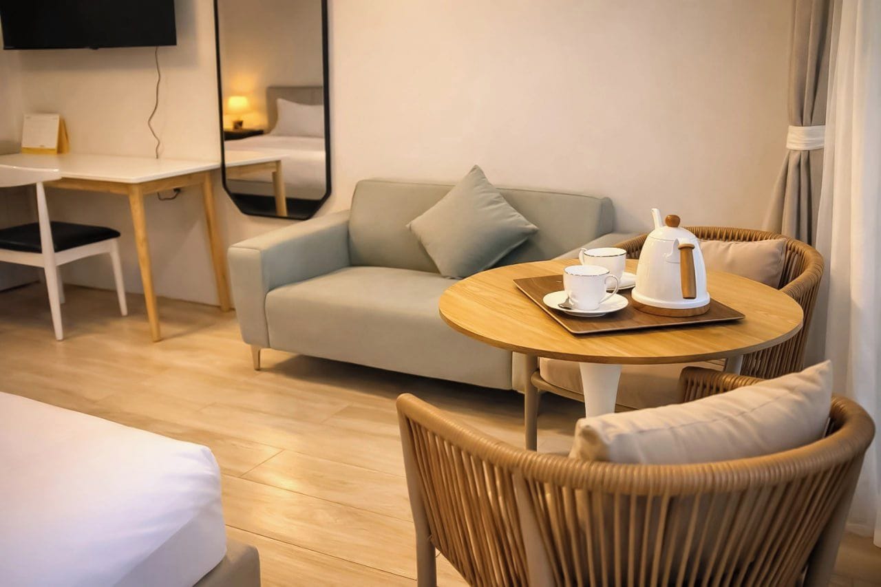 A hotel room with a bed, a sofa, a round dining table with a kettle and cups, a TV mounted on the wall, and a wooden floor.