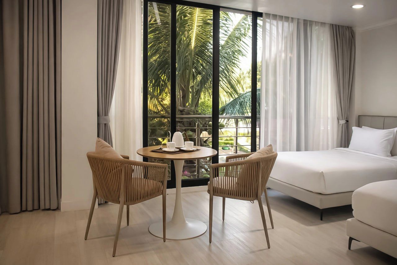 Een hotelkamer met een bed, twee stoelen, een klein rond tafeltje met een kopje en schotel, en uitzicht op palmbomen door grote ramen.