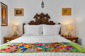 Un lit recouvert d’une couette blanche et d’une couverture à motifs floraux colorés, avec deux tables de chevet de chaque côté, sur lesquelles sont posées des lampes et des vases décoratifs.