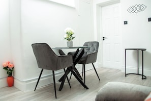 A modern dining area with a glass table, two grey upholstered chairs, and a potted plant. - Central Wembley - Apartment - Sleeps 5 - Parking (Wembley)