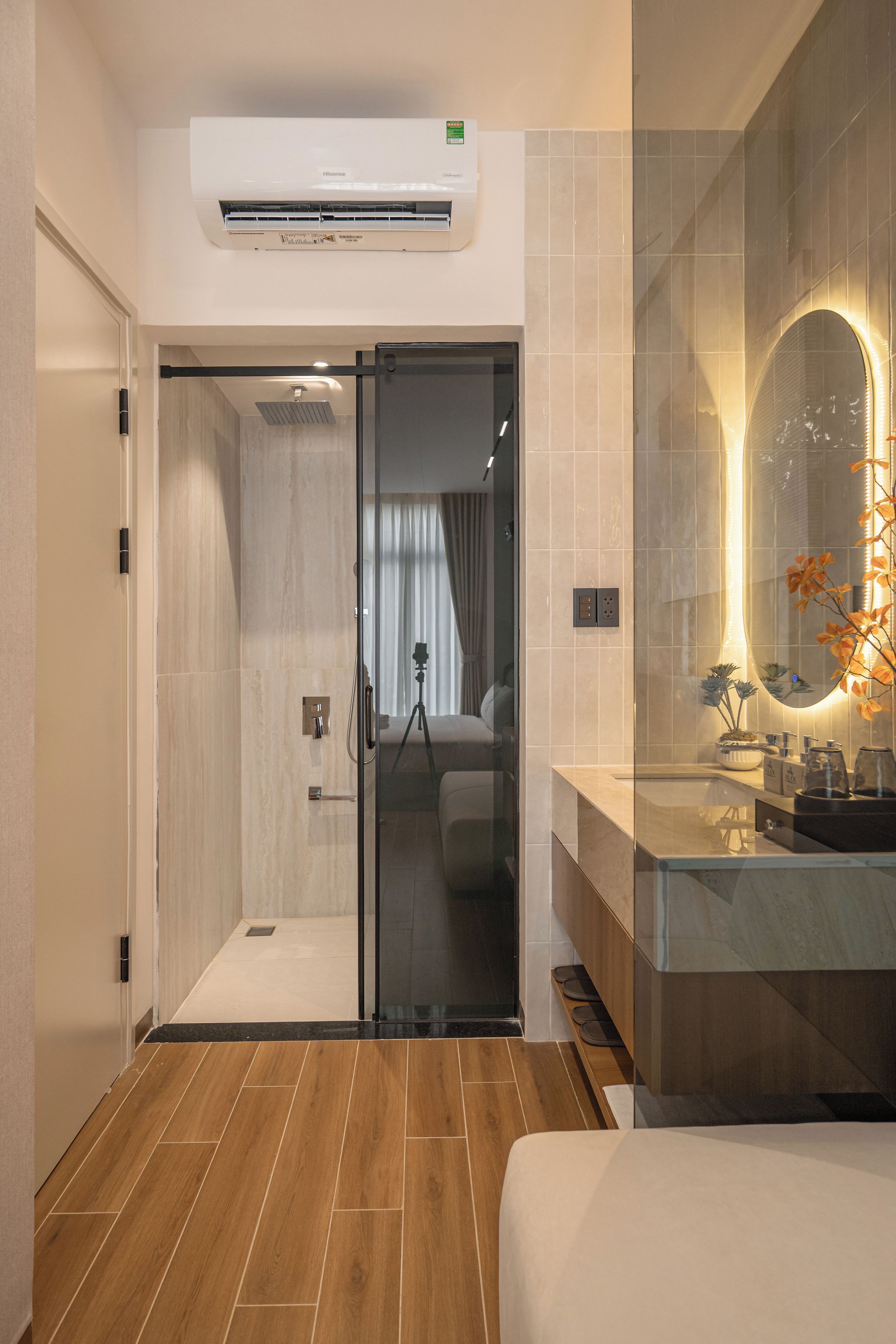Un baño moderno con cabina de ducha de vidrio, tocador con espejo y una cama en la habitación contigua.