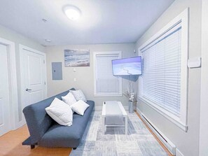 A modern living room with a grey sofa, a flat-screen TV mounted on the wall, and a window with blinds. - V-2BR Laneway - 15 Mins To DownTown (Vancouver)