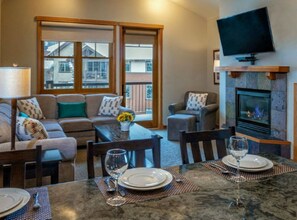 A living room with a fireplace, a television mounted above it, a sofa set, a dining table with chairs, and a view through large windows. - Grand Timber Lodge 1bd, Hot Tubs, Pools & Saunas (Breckenridge)