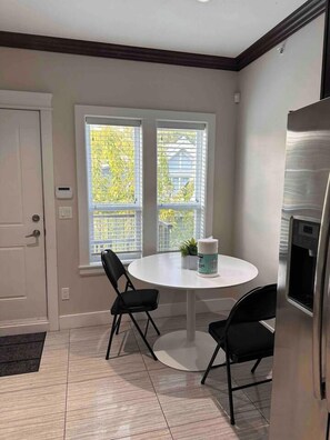 A kitchen with a round table, two chairs, a refrigerator, and a window with blinds. - V-2Spaces one area-near PNE (Vancouver)