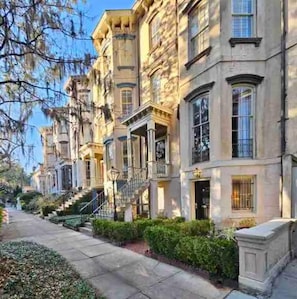 A row of townhouses with a central staircase, a sidewalk, and a clear sky. - Grand 1882 Victorian with 5BR, Steps to Forsyth Park (Savannah)