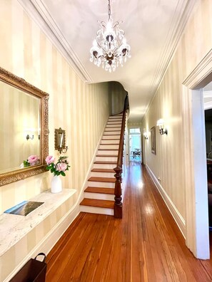 Un pasillo con una escalera de madera, un candelabro, un espejo y un florero con flores.