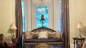 A room with a large chandelier, a sofa set, a dining table, and a view through a window.