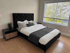 A modern bedroom with a black upholstered bed, a nightstand with a drawer, a potted plant, and a window with blinds. - Spacious 3bdrms in Vancouver for 8 people. NearPNE (Vancouver)