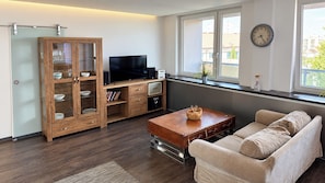 Una sala de estar moderna con un sofá, un mueble de madera con puertas de cristal, un televisor de pantalla plana y una ventana con vistas a edificios.