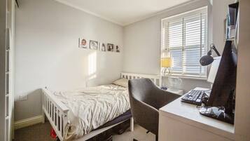 A bedroom with a single bed, a desk with a computer, a chair, and a window with blinds.