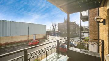 A balcony with a view of a street, a building with a metal fence, and a brick wall.