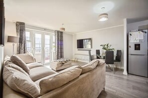 A living room with a sofa, a coffee table, a dining table with chairs, a TV, and a refrigerator. - Apartment Sleeps 4 with Balcony (Erith)
