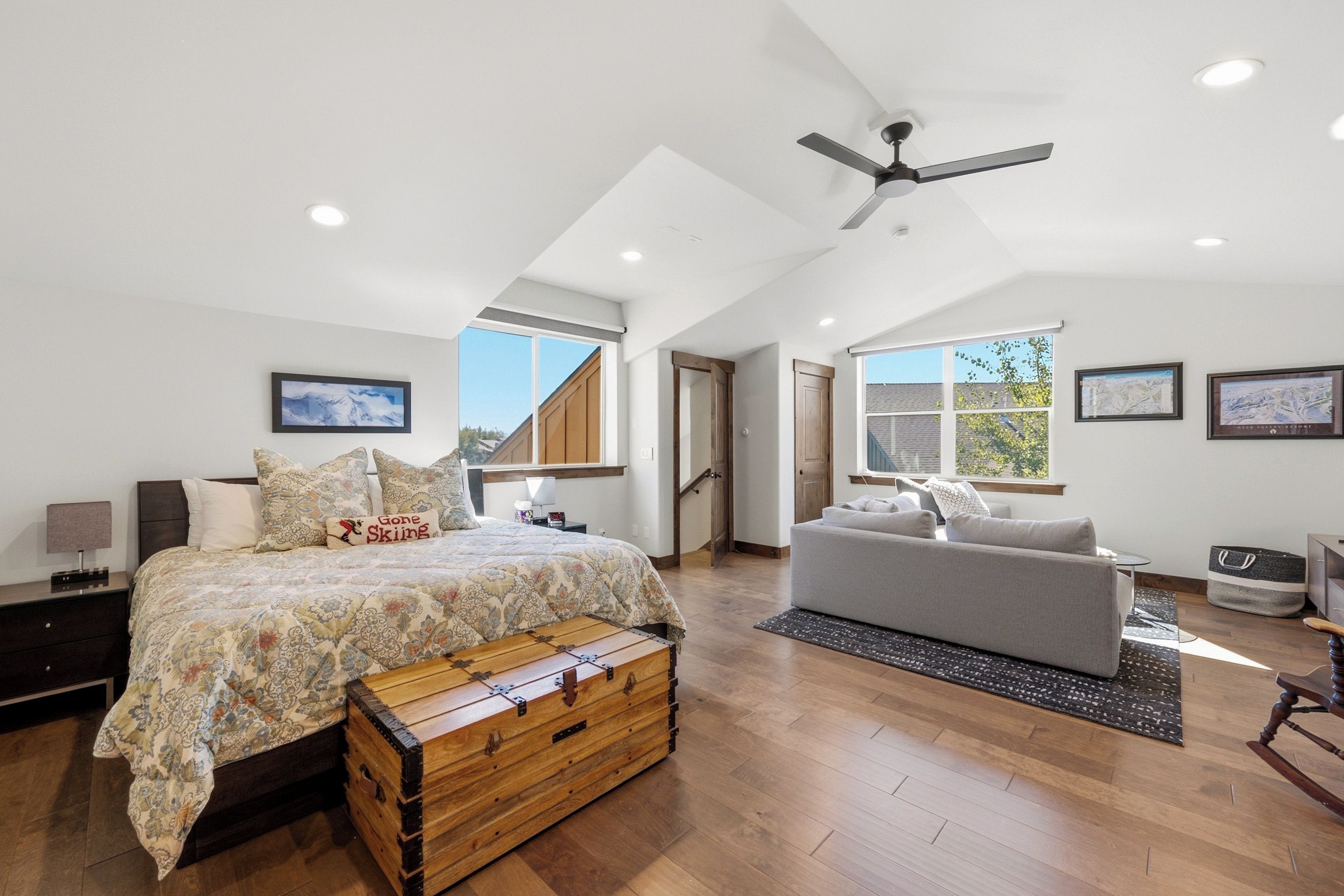 A bedroom with a bed, a chest of drawers, a grey sofa, a wooden chest, a ceiling fan, and large windows.