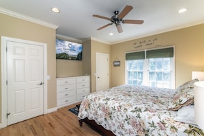 A bedroom with a bed, a ceiling fan, a window with blinds, a door, and a wall-mounted TV displaying a vacation rental sign. - Cheehaw Paradise | 2 Min Beach Walk | Elevator | Dual King Suites | Sleeps 14 | Dog Friendly (Edisto Beach)