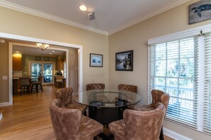 A dining area with a glass table surrounded by four chairs, a kitchen in the background, and a window with blinds. - Cheehaw Paradise | 2 Min Beach Walk | Elevator | Dual King Suites | Sleeps 14 | Dog Friendly (Edisto Beach)