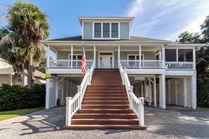 A two-story house with a front porch, a staircase leading to the entrance, and a flag on the railing. - Cheehaw Paradise | 2 Min Beach Walk | Elevator | Dual King Suites | Sleeps 14 | Dog Friendly (Edisto Beach)