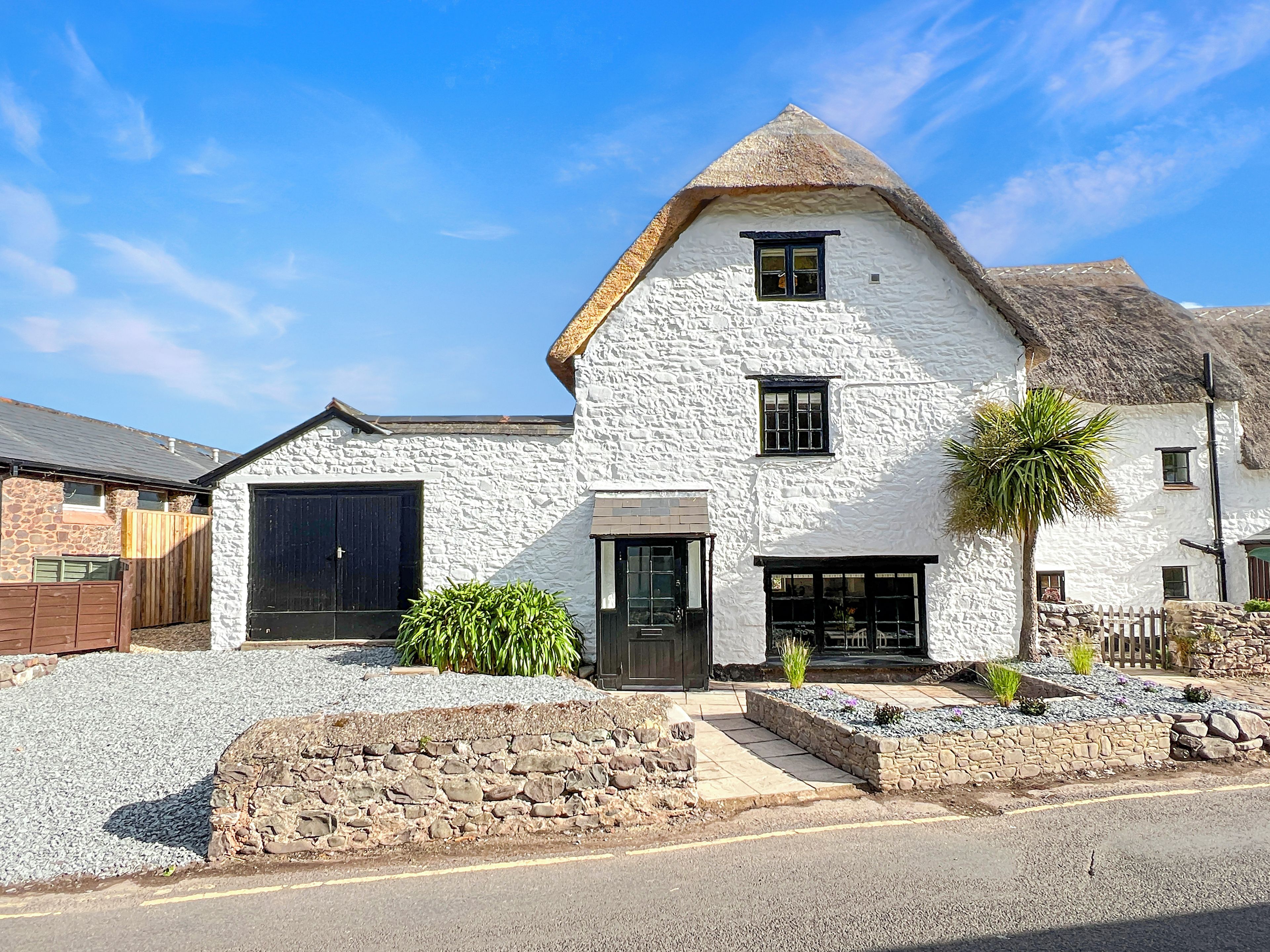 A white stone house with a thatched roof, a black garage door, and a small garden with gravel and plants.
