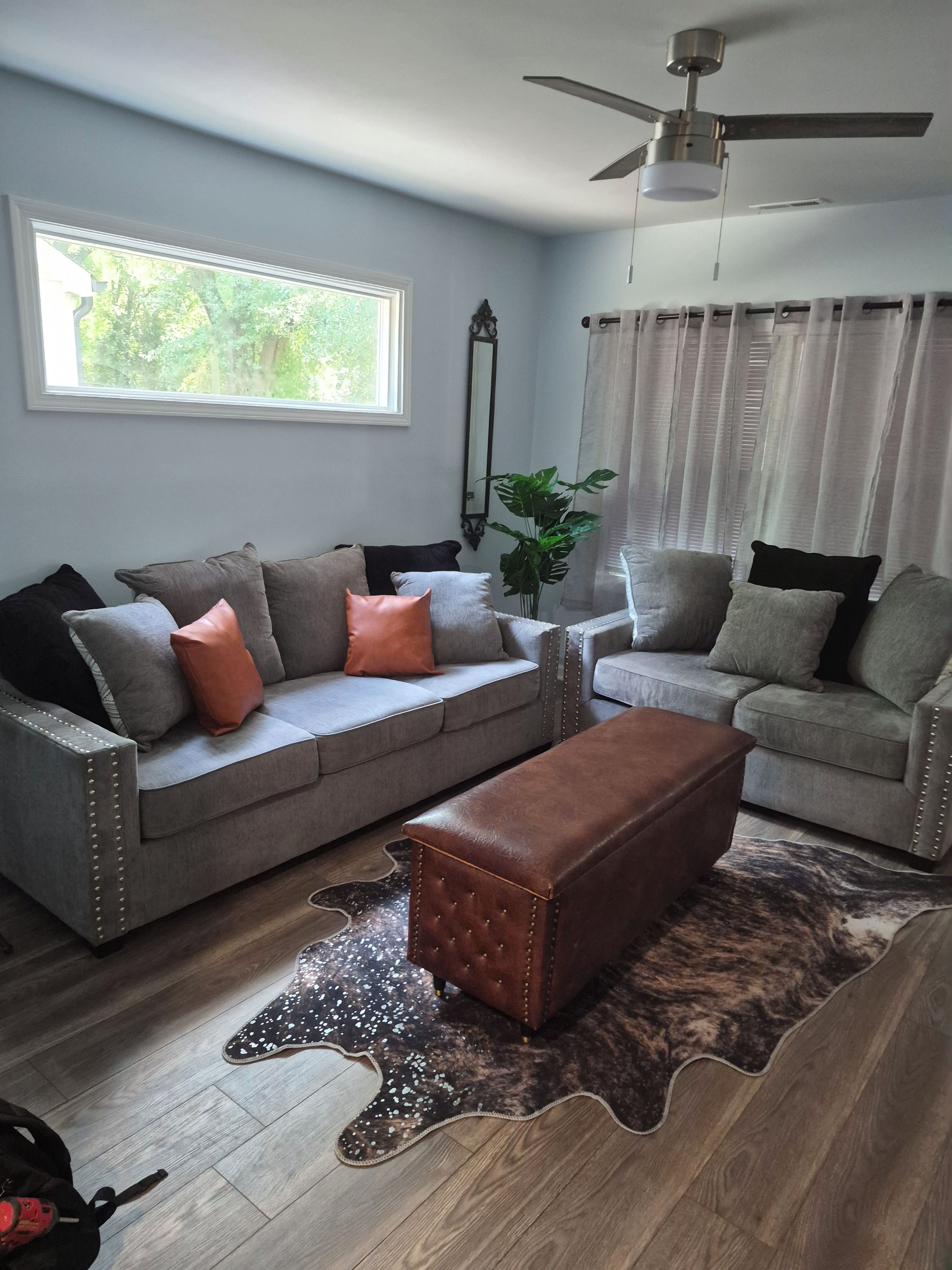 A living room with a grey sofa, a brown ottoman, and a ceiling fan.