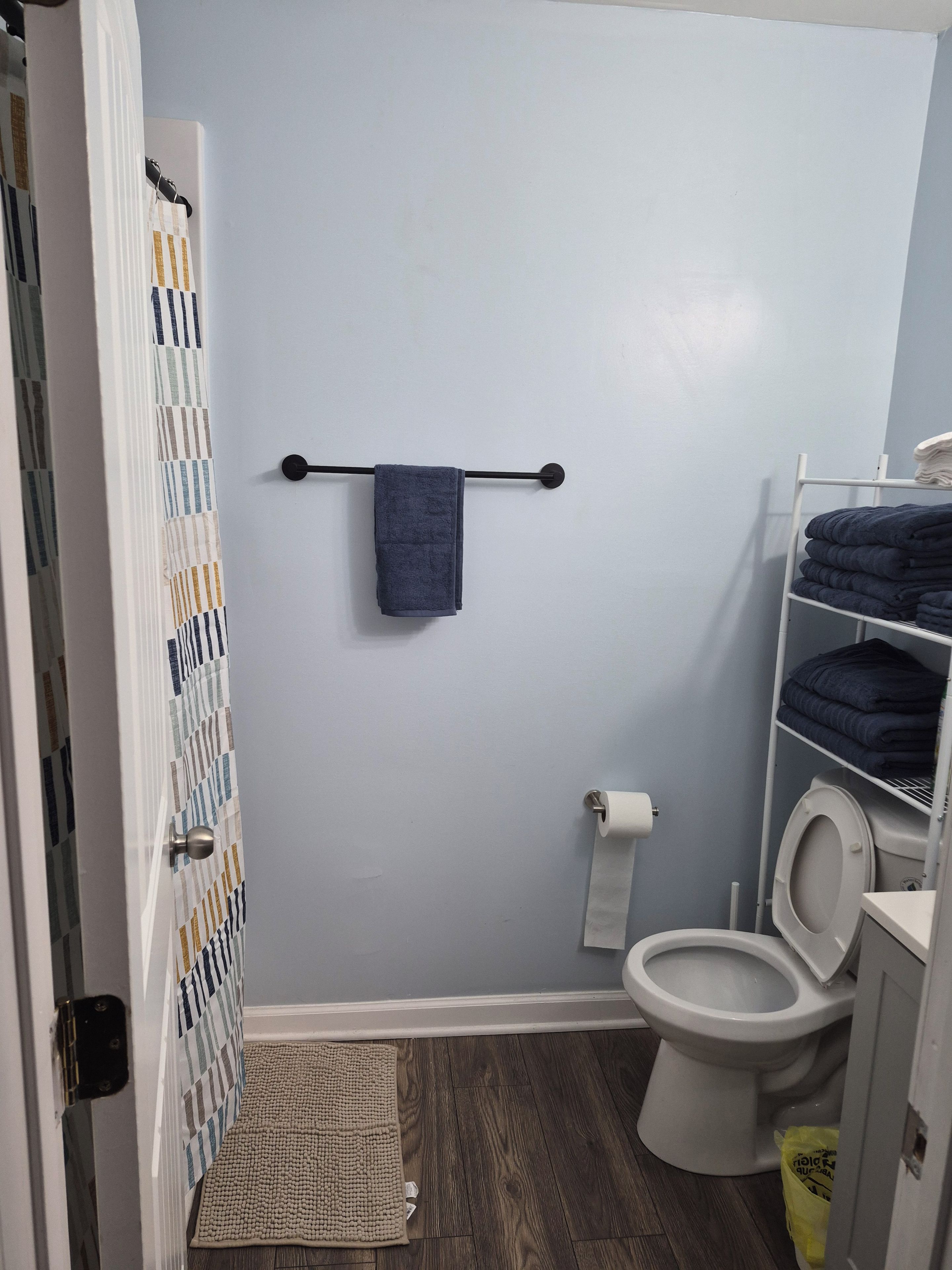 A bathroom with a toilet, a towel rack with a towel, a toilet paper holder, a shelf with folded towels, and a patterned shower curtain.
