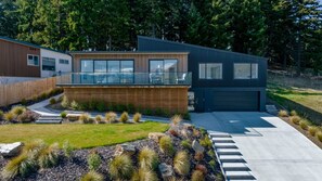 A modern two-story house with a garage, a covered patio, and a landscaped garden with native plants. - Elevated Lake Wānaka Retreat | 4 Bedrooms, 3 Bathrooms, Fireplace & Panoramic Views (Wānaka)