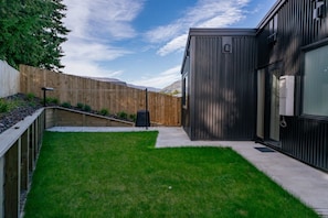 A modern building with a dark exterior, a grassy yard, a wooden fence, and a clear sky. - Elevated Lake Wānaka Retreat | 4 Bedrooms, 3 Bathrooms, Fireplace & Panoramic Views (Wānaka)