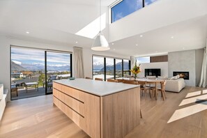 A modern kitchen with a large island, dining area, and a TV. - Elevated Lake Wānaka Retreat | 4 Bedrooms, 3 Bathrooms, Fireplace & Panoramic Views (Wānaka)