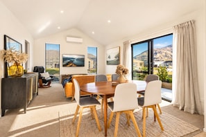 A modern dining area with a wooden table and chairs, a sofa, and a view of a mountainous landscape through large windows. - Modern Wānaka Retreat | 3 Queen Bedrooms, Mountain Views, High Ceilings & Internal Garage (Wānaka)