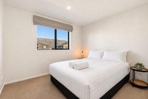 A hotel room with a large bed, a bedside table with a lamp, a window with a view of buildings, and a small plant on the nightstand. - Modern Wānaka Retreat | 3 Queen Bedrooms, Mountain Views, High Ceilings & Internal Garage (Wānaka)